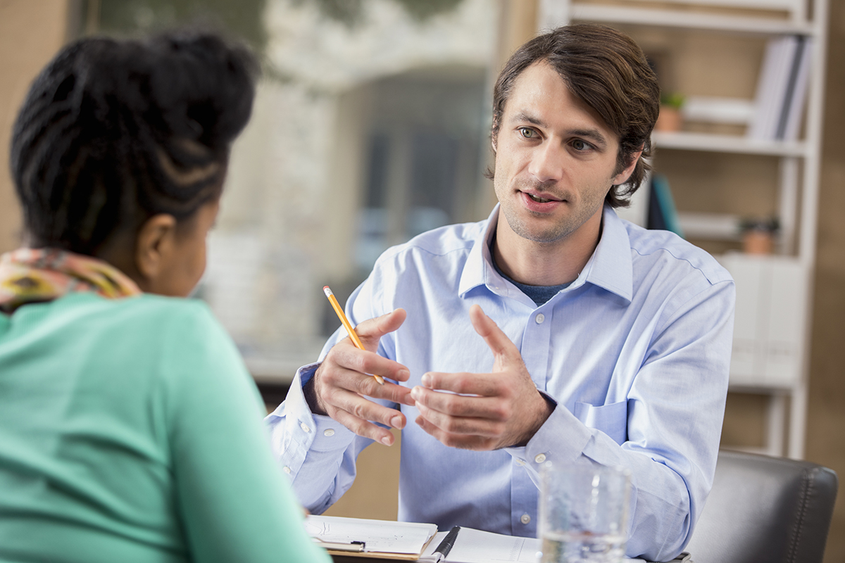 Master of Business Administration students talking, one, a man with brown hair, facing towards the camera using his hand to gesture and the other, a woman wearing an aqua green long sleeved top, looking at him, away from the camera.